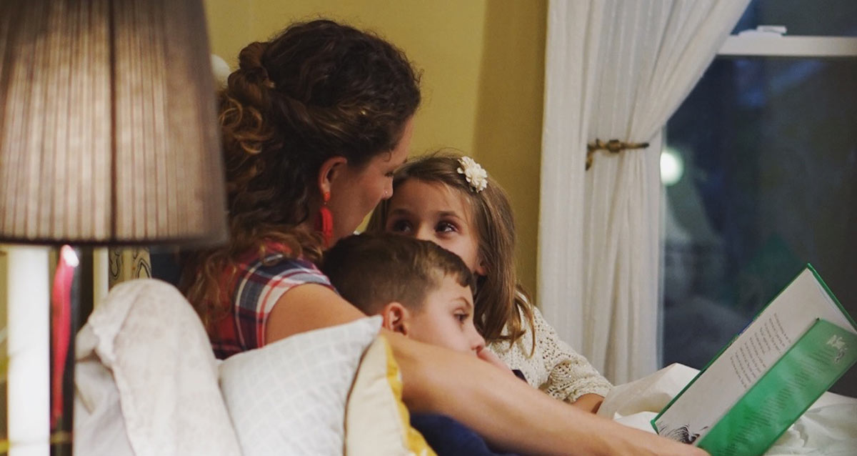 how to build child bedtime routine, Image contains a photo of a white mother and two young kids (a boy and a girl) in bed reading a storybook.