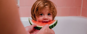 Image contains a photo of a child eating a slice of watermelon in the bathtub as they develop a healthy habit at a young age.