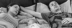 Image contains two new parents sleeping on a bed with a newborn between them.