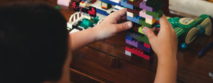 Image contains a photo of a child playing with blocks on a woodern surface to develop their physical and mental skills.
