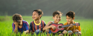 Image contains a photo of four children sitting in long grass playing a small guitar.
