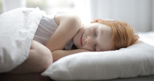 best ways to get child to sleep, Image contains a photo of a red-haired girl laying in her bed with her head on a pillow. Her eyes are closed and her knee is visible just above the blanket on top of her.